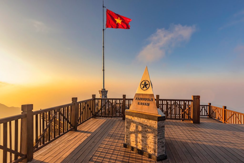 View from the summit of Fansipan mountain in Vietnam, featuring a flagpole with the Vietnamese flag and a stone monument marking the peak at 3,147 meters.