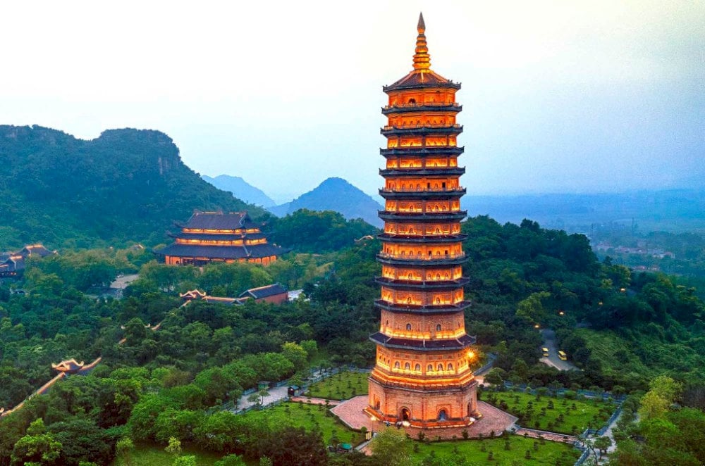 A scenic view of a tall, illuminated pagoda surrounded by lush greenery and mountains during dusk.