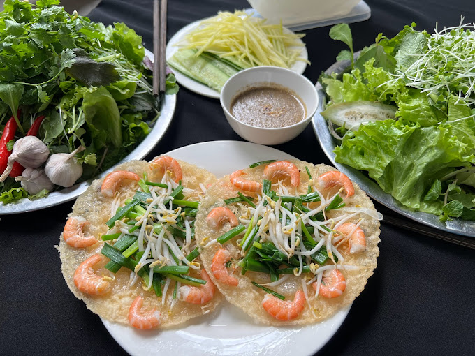 A plate of two shrimp pancakes topped with green onions and bean sprouts, surrounded by fresh herbs, lettuce, cucumbers, and a small bowl of dipping sauce.