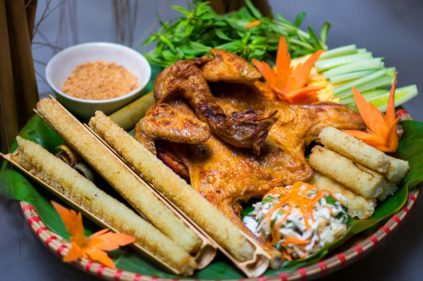 A vibrant platter of Vietnamese cuisine featuring grilled chicken, traditional bamboo rice rolls, fresh vegetables, and a small bowl of spicy dipping powder. com lam