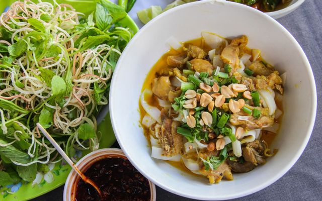 A bowl of noodle soup topped with meat, green onions, and peanuts, surrounded by fresh herbs and a side of spicy sauce.