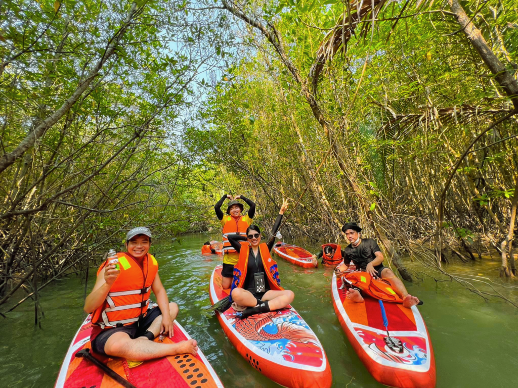 sup-can-gio-forest Four people wearing orange life jackets sitting on paddleboards in a narrow waterway surrounded by dense green mangrove trees