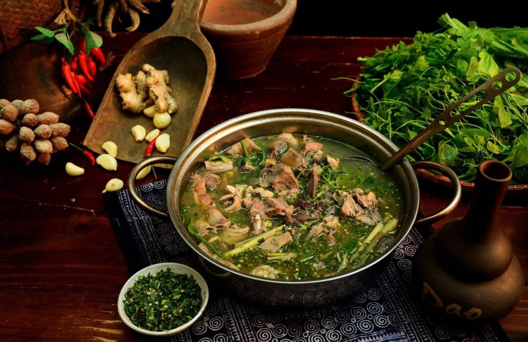 A pot of hearty soup with meat and herbs, surrounded by fresh ingredients including ginger, garlic, chili peppers, and green vegetables.