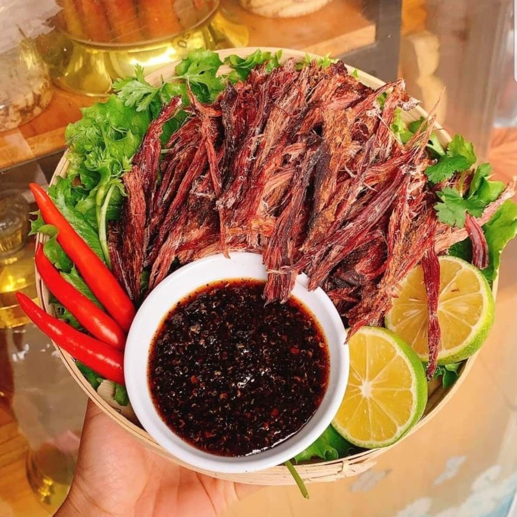 A woven basket filled with dried meat, fresh greens, red chili peppers, lime slices, and a small bowl of dark dipping sauce.