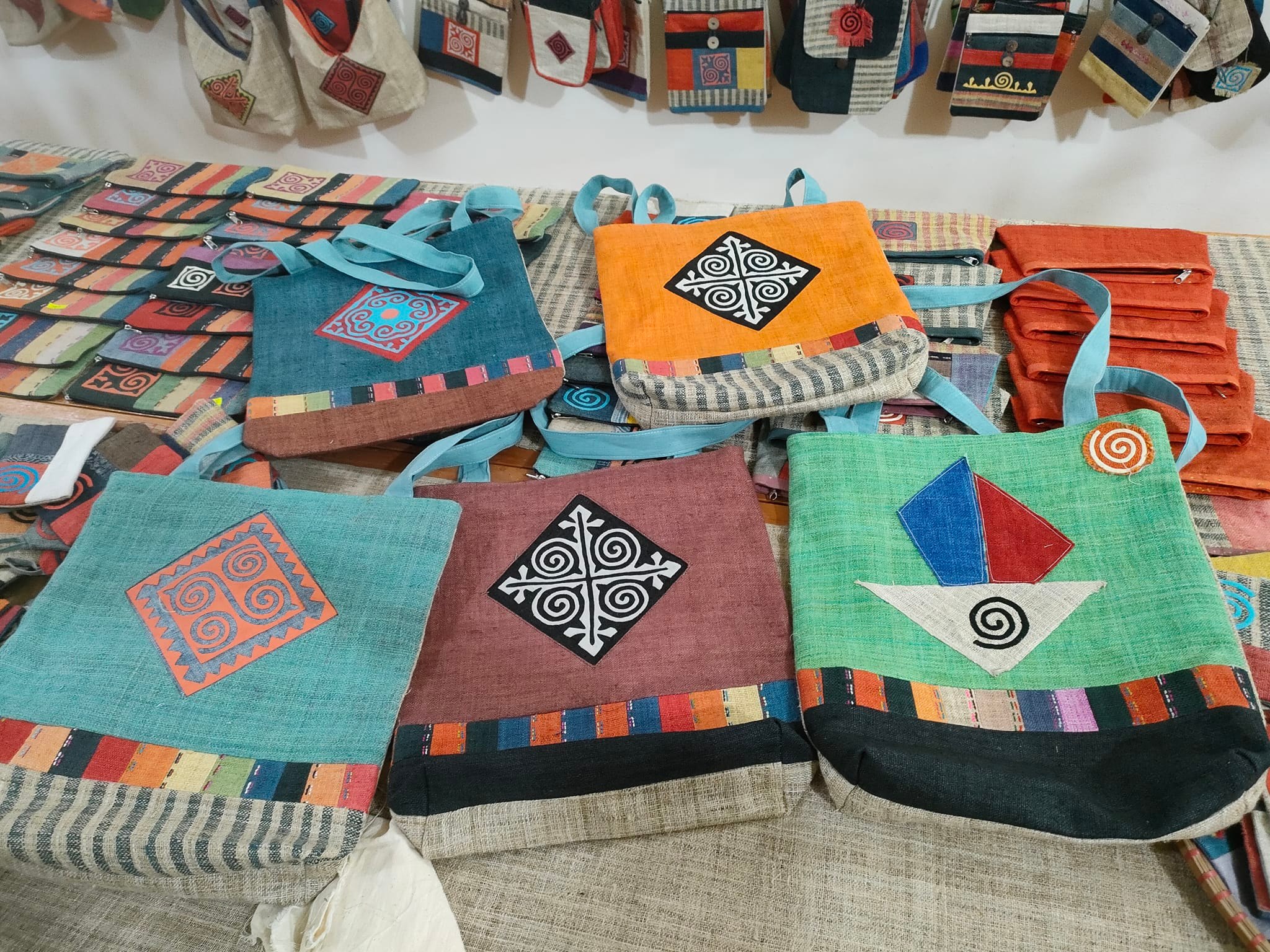 A collection of colorful handmade bags displayed on a table, featuring various patterns and designs. The bags are primarily in shades of turquoise, orange, and maroon, with decorative motifs and straps. Behind them, smaller bags are hung, showcasing similar vibrant colors and designs.