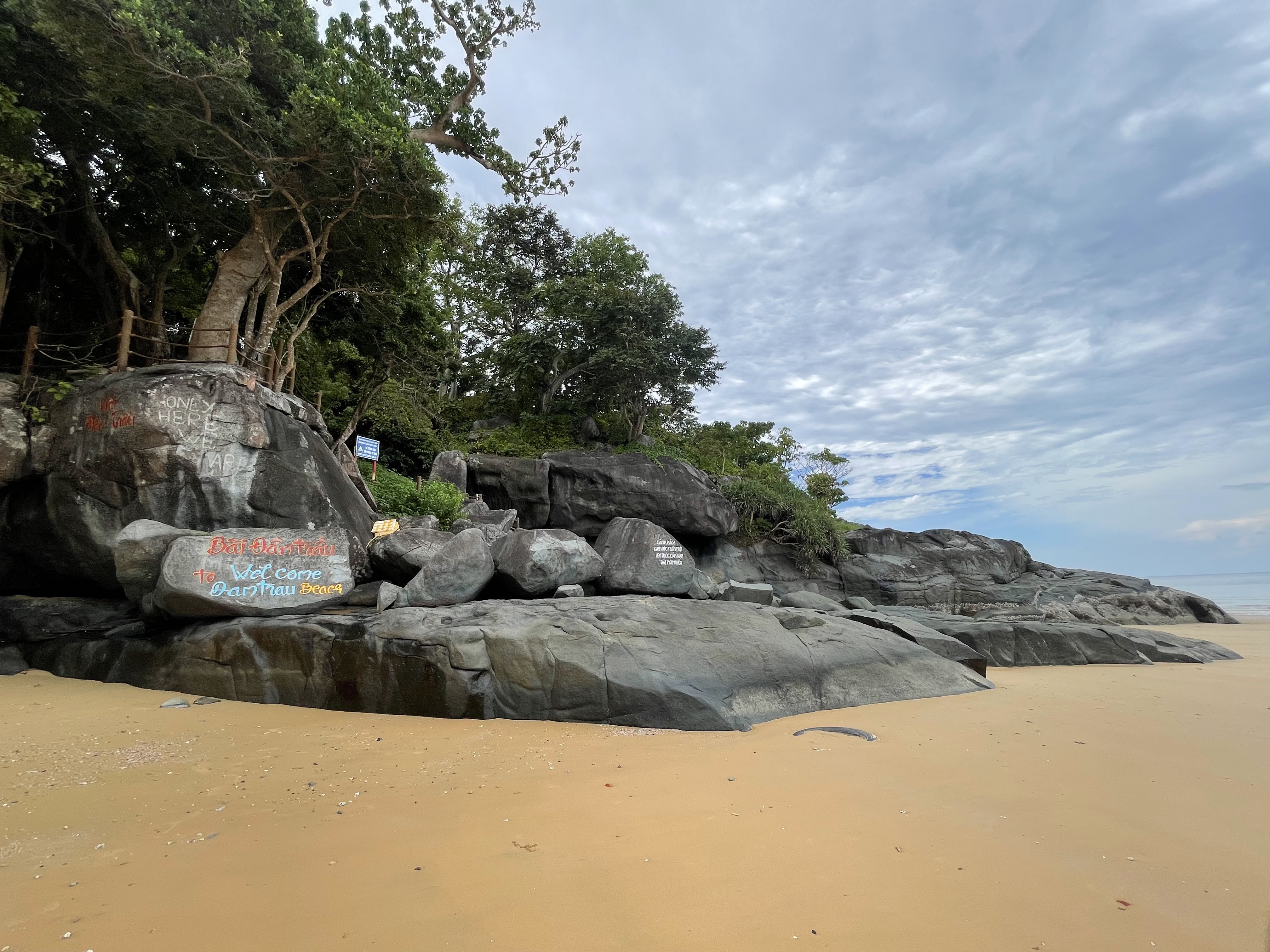 Rocky beach area with large boulders, greenery, and a cloudy sky.