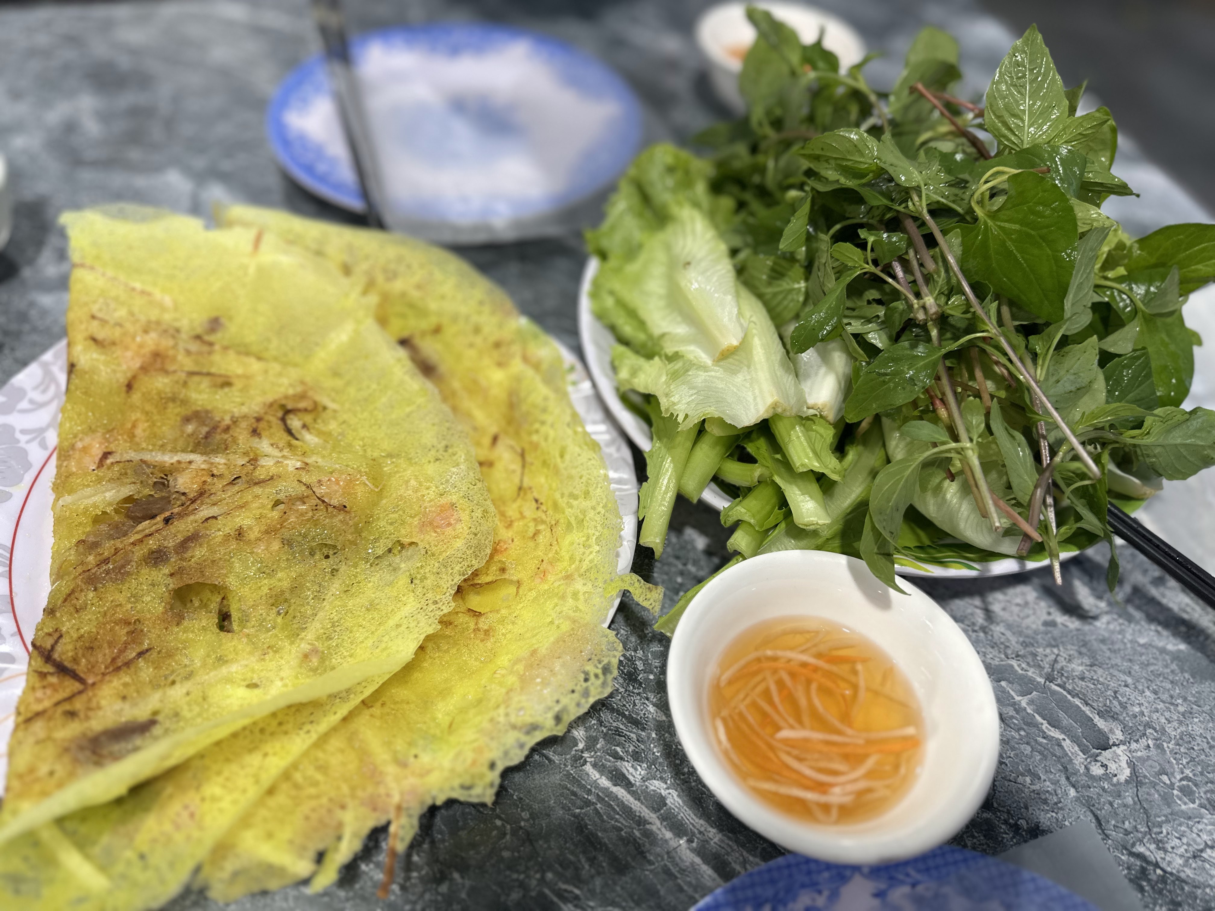A plate of golden Vietnamese pancakes topped with shrimp and herbs, accompanied by a side of fresh greens and a bowl of dipping sauce.