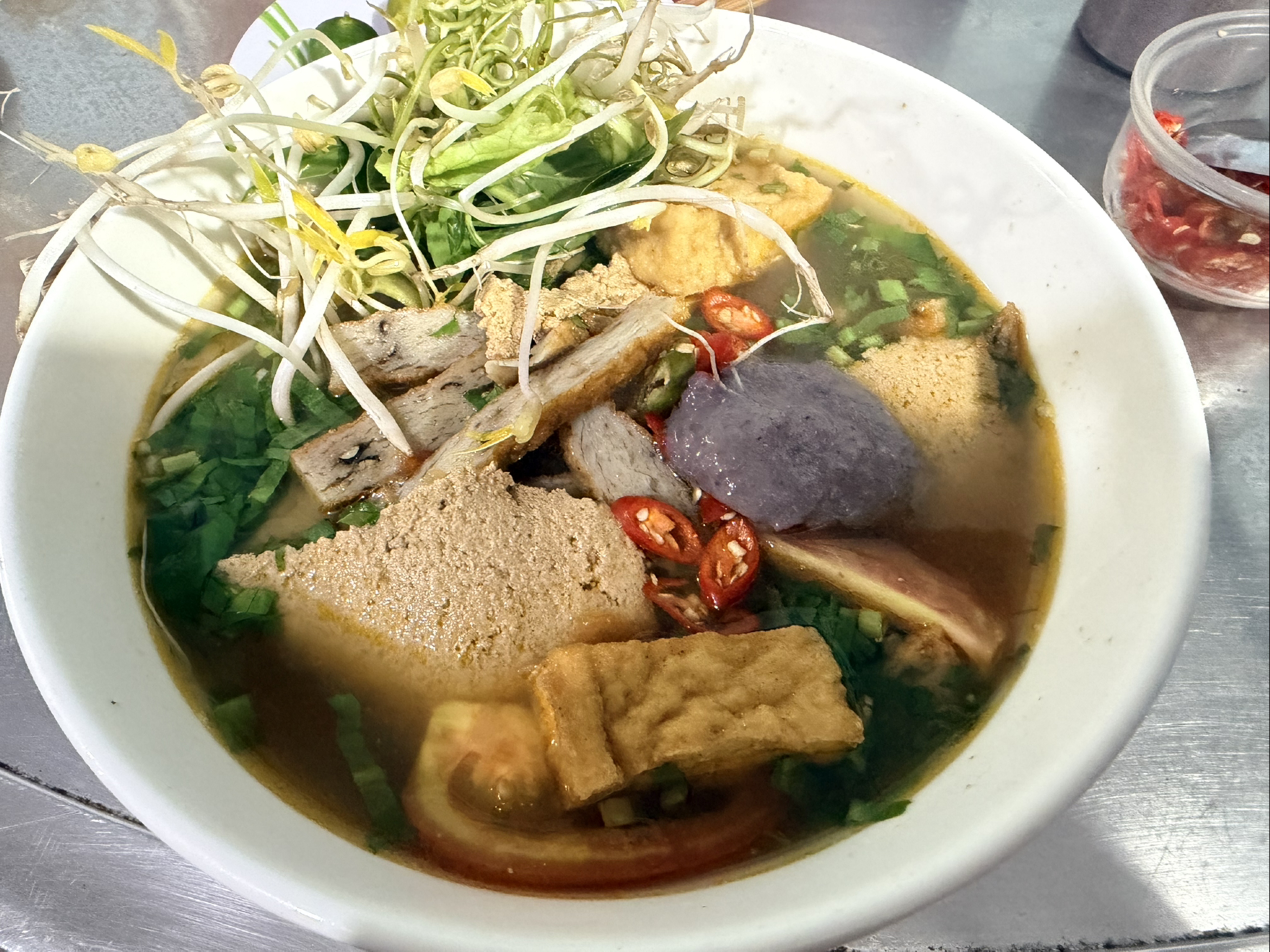 A bowl of noodle soup featuring various ingredients such as noodles, herbs, slices of meat, tofu, and chili peppers, garnished with bean sprouts and greens.