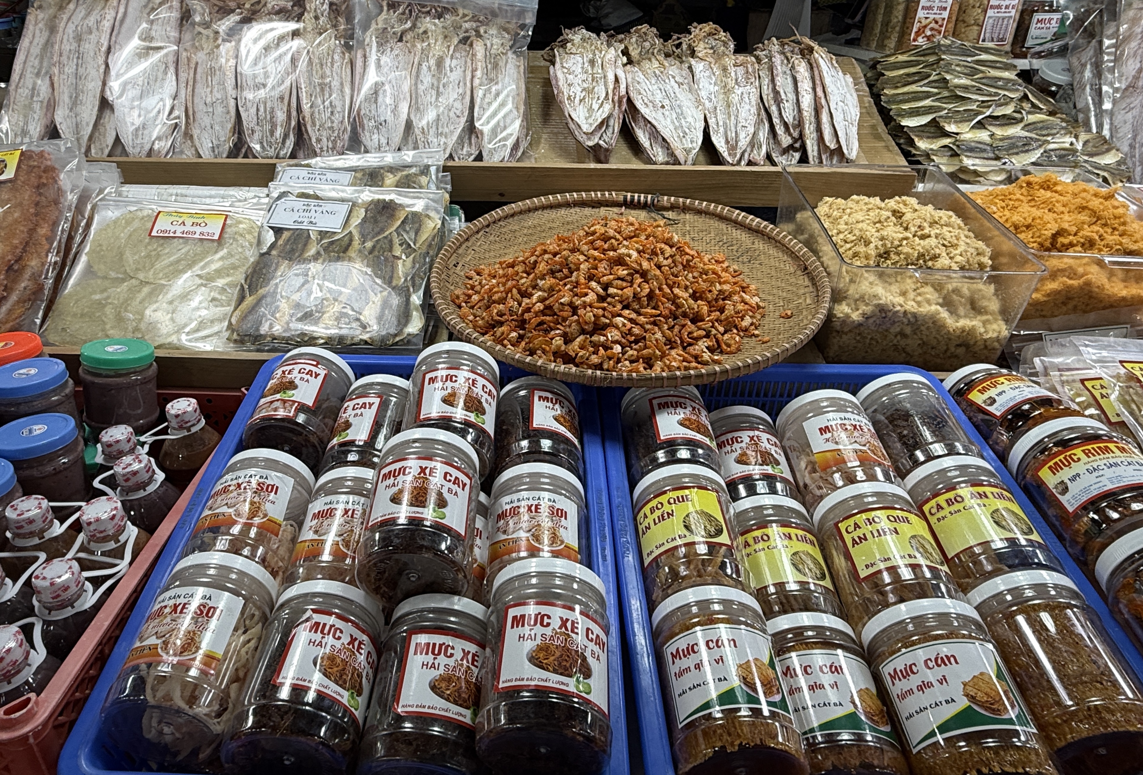 A vibrant market display featuring a variety of dried seafood, spices in jars, and a woven basket of small dried shrimp. The table is filled with colorful packages and labels in Vietnamese.