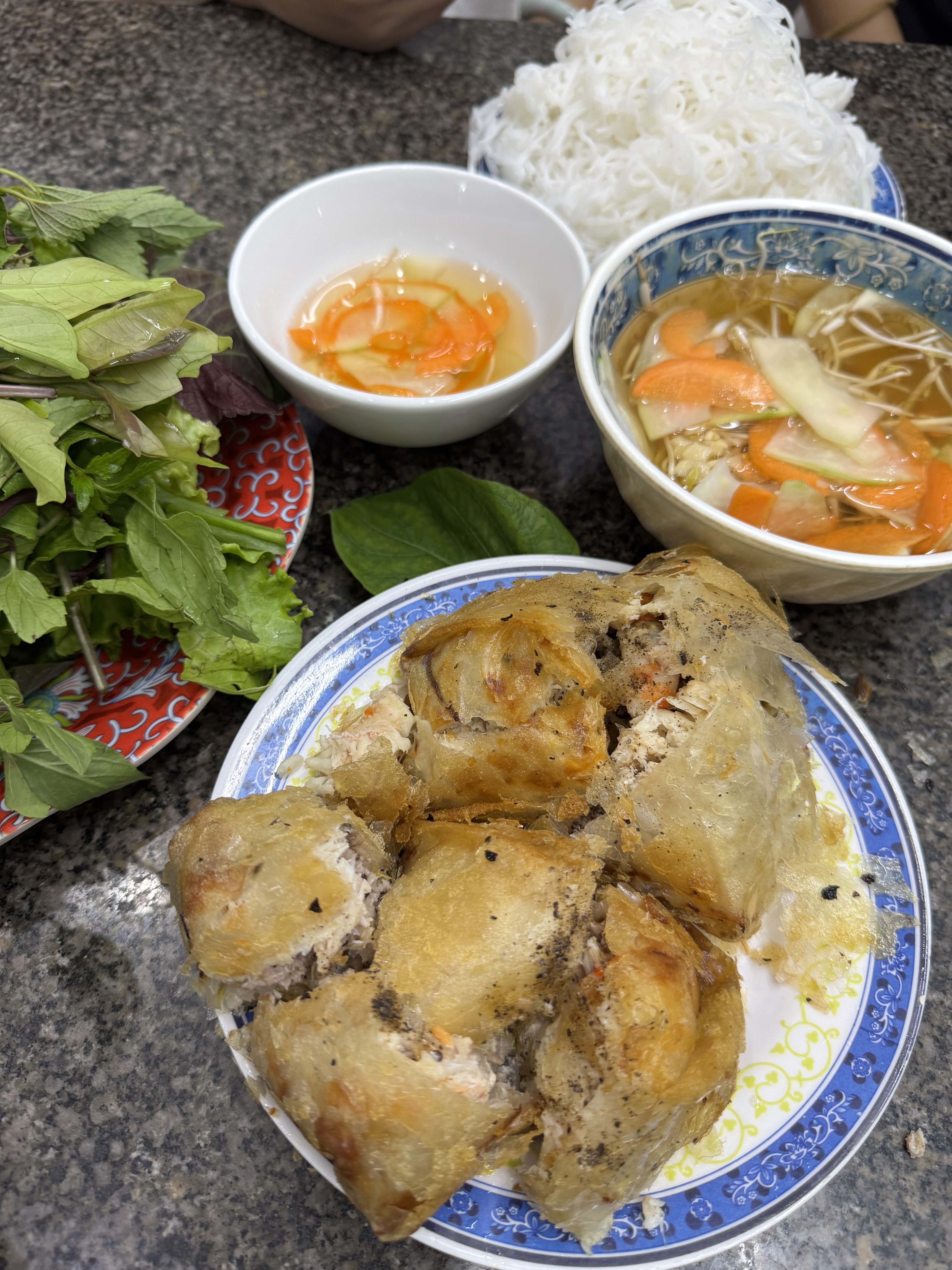 A plate of fried spring rolls with a side of fresh herbs, a bowl of dipping sauce, and a bowl of noodle soup with vegetables, served on a granite countertop.