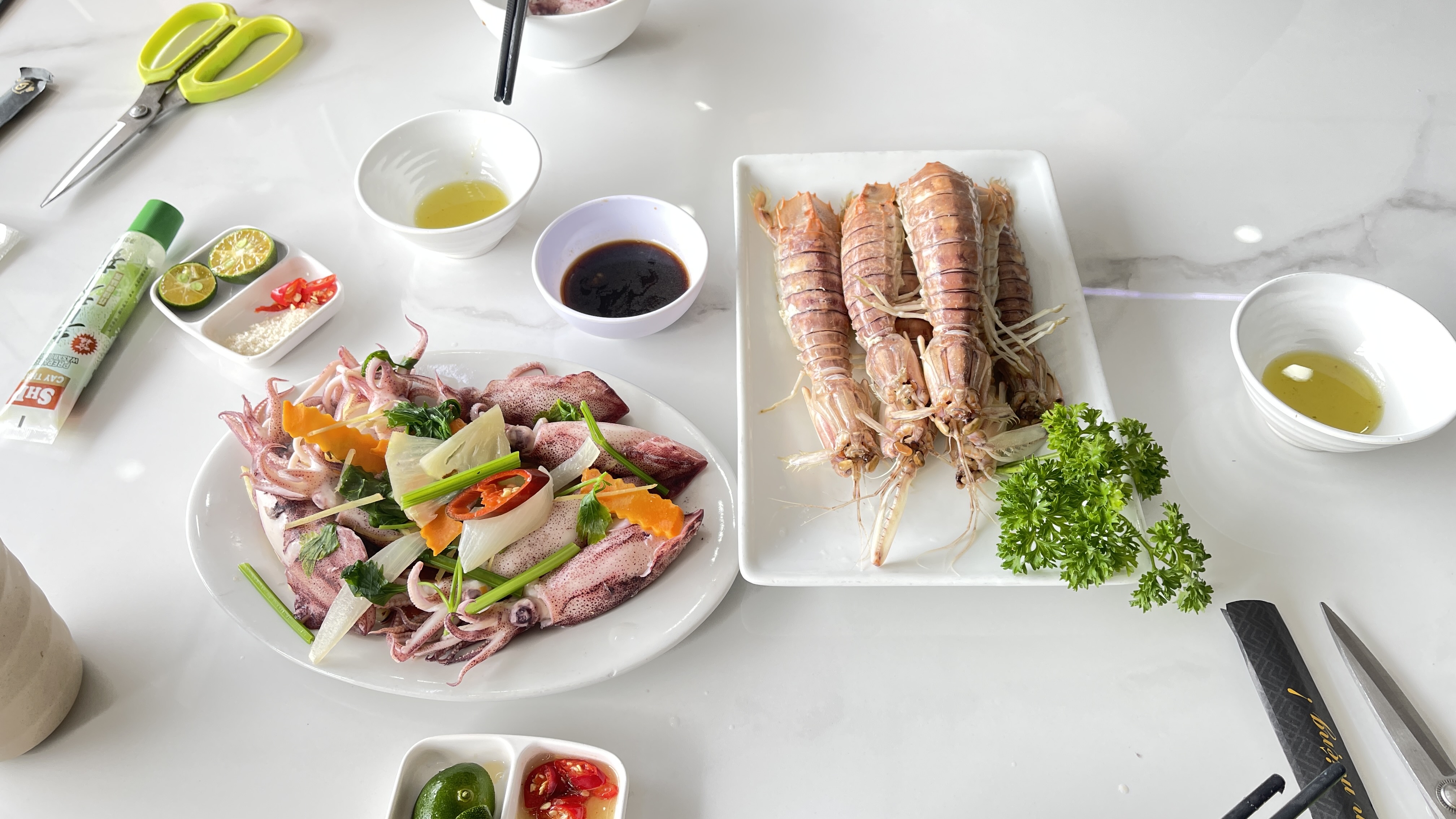 A table setting featuring a plate of fresh squid garnished with herbs and vegetables, alongside a platter of large shrimp. Various dipping sauces, lime wedges, and sliced chili are arranged neatly on the table, with kitchen scissors and utensils visible in the background.