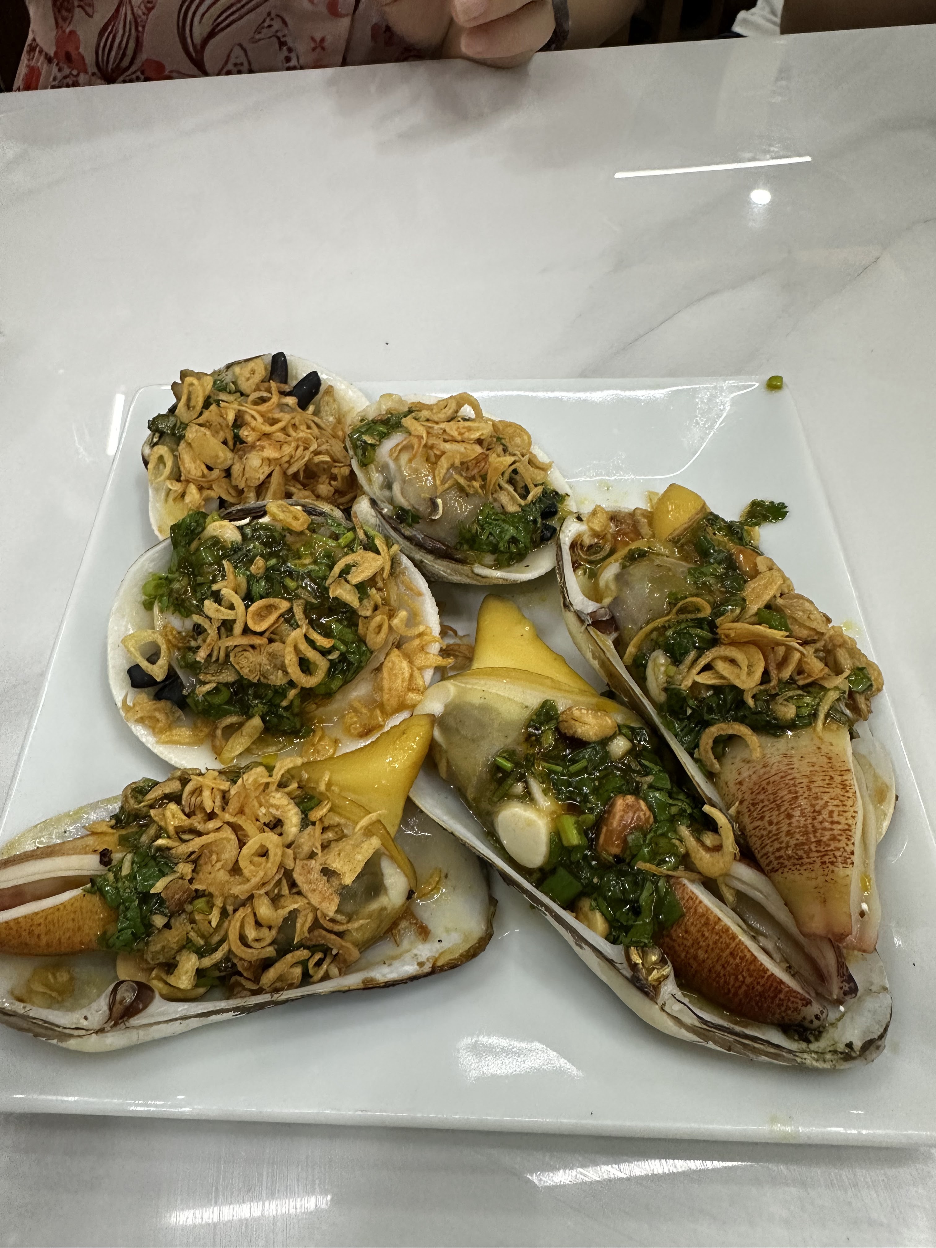 A plate of seafood featuring several large, grilled clams topped with green herbs, crispy fried onions, and a vibrant sauce.