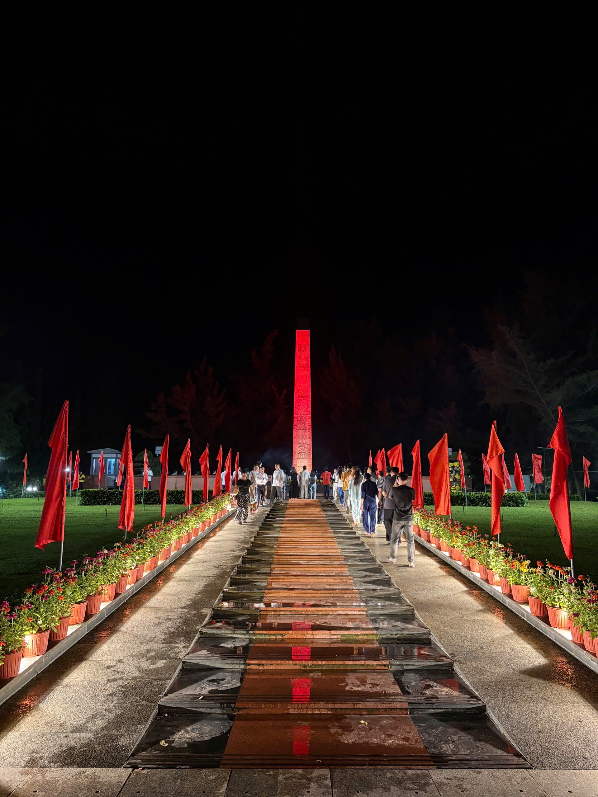 Pathway illuminated at night leading to a tall red monument, flanked by red flags and flower beds, with people walking towards it.