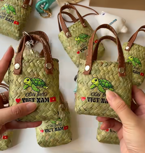 A close-up of small woven bags featuring a green turtle design and the text 'CON ĐẢO VIET NAM', held by a person with two bags visible in the foreground.